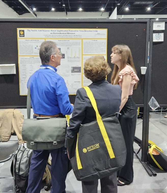 Three individuals at a conference, with one presenting research findings on a poster to the other two attendees.