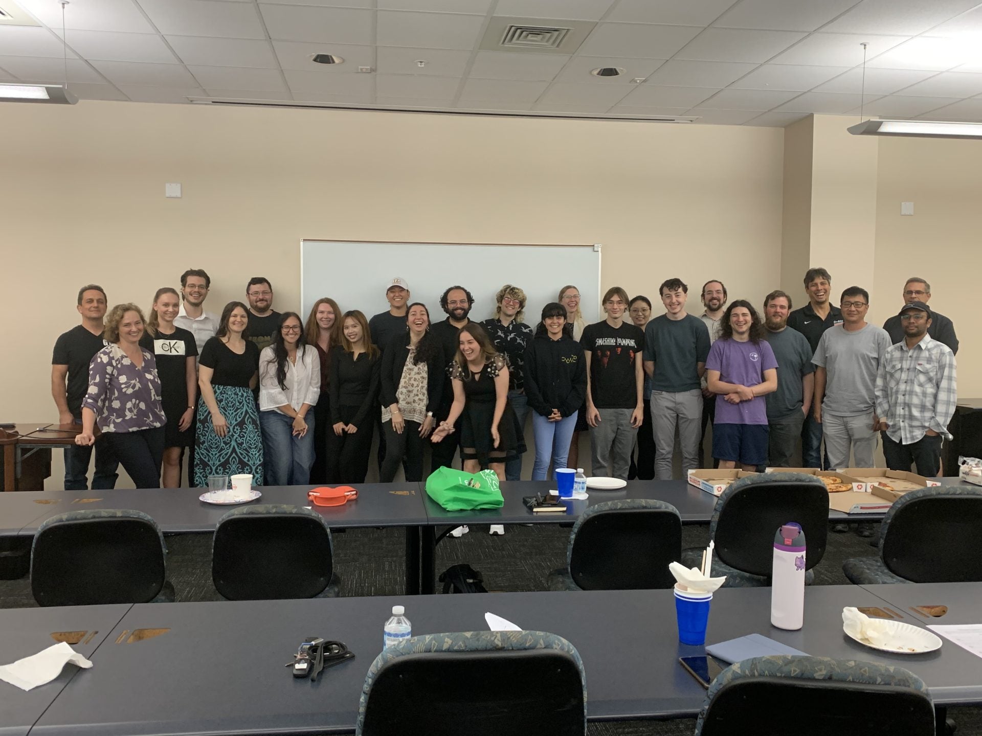 A group of people stand together in a classroom-like setting, smiling for a photo. There are tables with food and drinks in front of them.