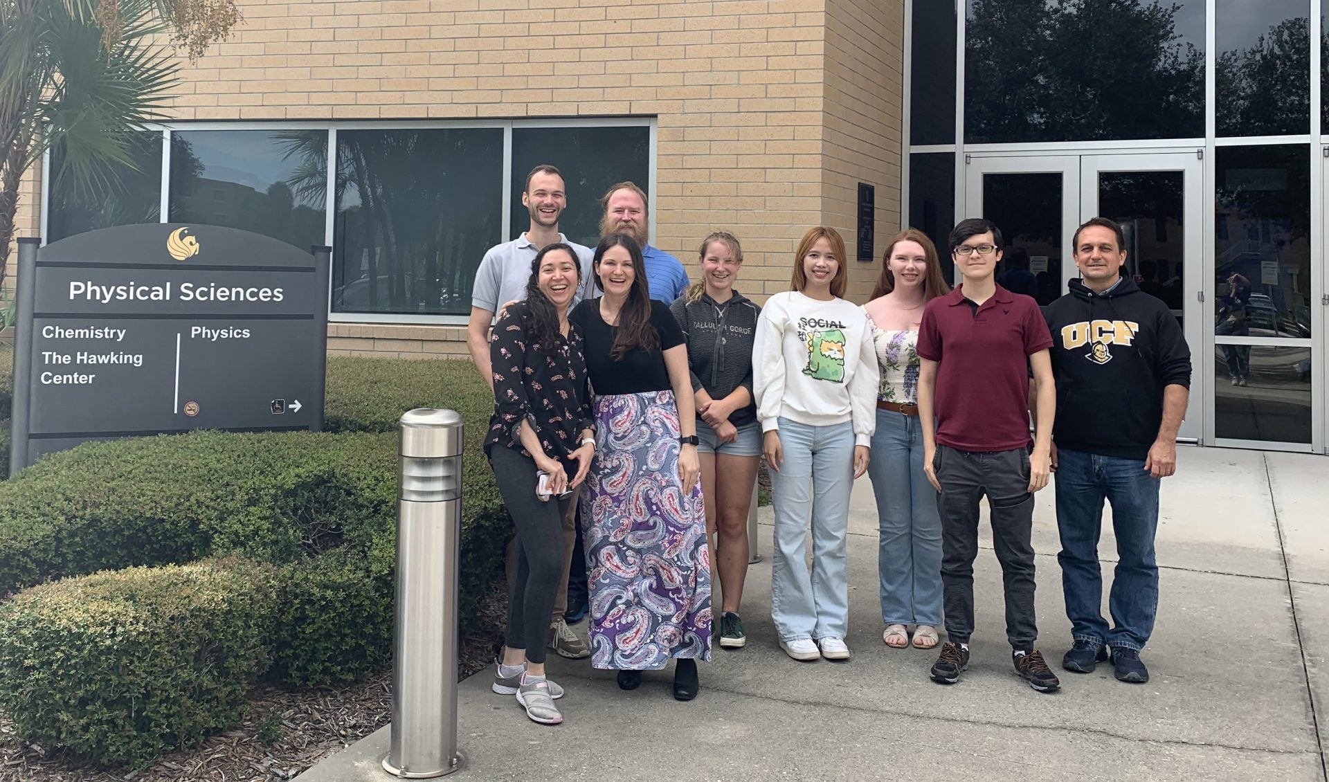 A group of nine people stand smiling outside a building next to a sign that reads "Physical Sciences, Chemistry, Physics, The Hawking Center.
