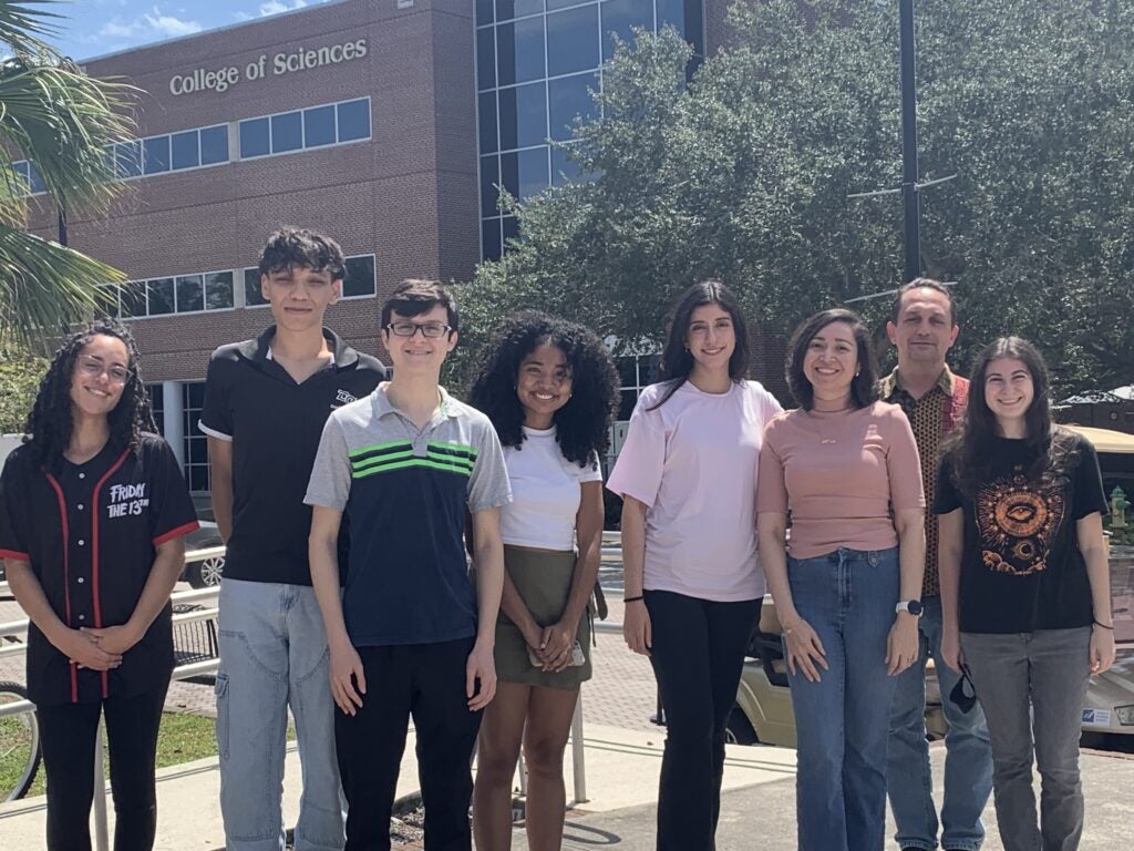 Eight people stand outside in front of a building labeled "College of Sciences" on a sunny day, posing for a group photo.