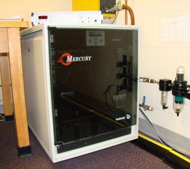 A Mercury Varian spectrometer in a laboratory setting, placed next to a wooden table with various connected tubing and equipment.